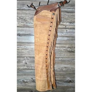 Puede incluir: Chaps de cuero color canela con flecos y un cinturón de cuero marrón. Los chaps tienen un bolsillo en la pierna izquierda.