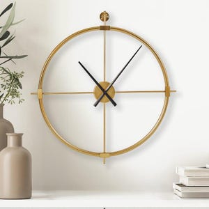 Op de afbeelding: Een goudkleurige, minimalistische wandklok met een cirkelvormig frame en zwarte wijzers. De klok heeft een eenvoudig ontwerp met een gouden midden en dwarsbalken, gemonteerd op een witte muur. De wijzers van de klok zijn zwart.
