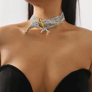 Peut inclure: Un collier ras du cou argenté et doré représentant un oiseau aux ailes déployées. Le collier est composé de plusieurs rangées de chaînes argentées. Le corps de l'oiseau est doré et les ailes sont argentées avec des pierres claires.