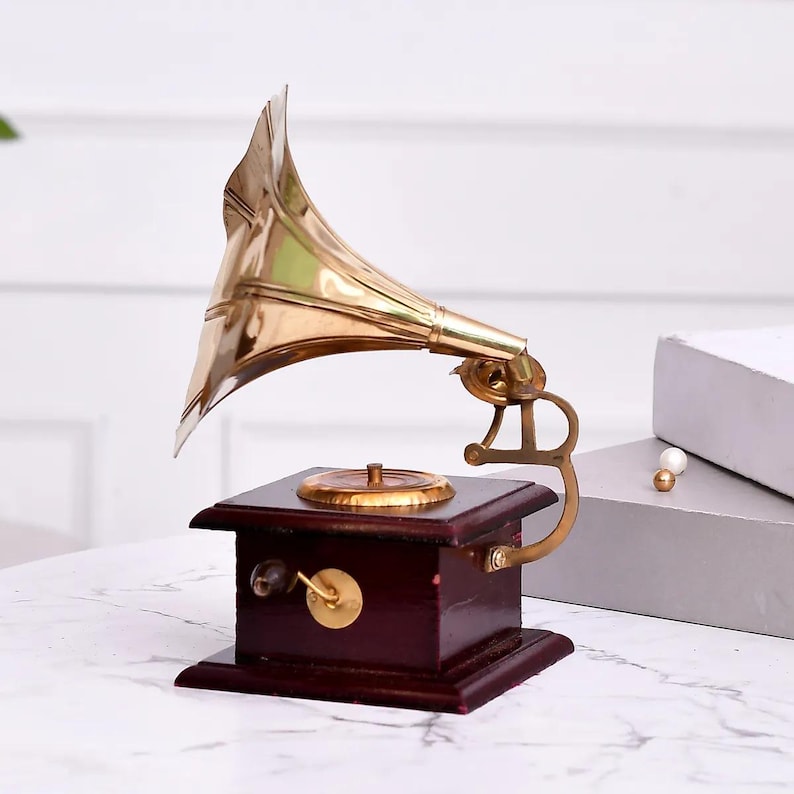 May include: A vintage-style gramophone with a polished brass horn and a dark wood base. The gramophone features a gold-colored turntable and a hand crank. This decorative piece evokes a classic, antique aesthetic.