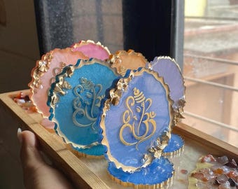 May include: A collection of resin coasters in blue, pink, and purple, each with a gold Ganesh design. The coasters have gold edging and are presented on a wooden tray with decorative stones. The coasters are approximately 10 cm in diameter.