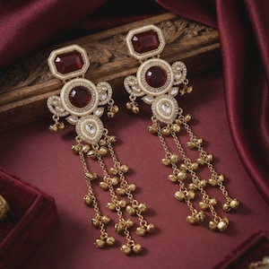 May include: A pair of ornate gold-toned earrings with ruby-red rectangular and circular gemstones. The earrings feature intricate detailing, including small dangling gold beads and teardrop-shaped accents. The background is a rich burgundy fabric.