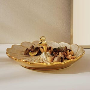 May include: A gold-colored decorative dish with a peacock-shaped handle. The dish has a leaf-like design and is filled with dried botanicals. The rim and peacock are a brighter gold color.