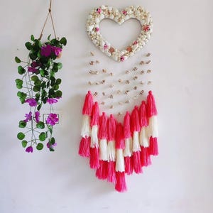 May include: A heart-shaped wall hanging made of white seashells and small pink flowers. Below, a cascade of pink and white tassels hangs from strings of small shells. To the left, a hanging plant with purple flowers.