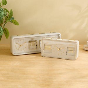 Pode incluir: Dois relógios analógicos retangulares creme com detalhes dourados. Os relógios exibem o dia e a data, com um mostrador transparente mostrando a hora. Estão em uma superfície de madeira.