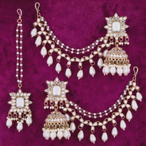 May include: An ornate gold-toned jewelry set featuring a headpiece and two earrings. Each piece has a square, mirrored center surrounded by a floral design. White teardrop-shaped pearls and delicate chains complete the design. The set is displayed on a deep purple background.