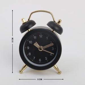 May include: A black alarm clock with gold-coloured accents. The clock face is black with gold hands and numbers. The clock is 5 cm wide and 8 cm tall.