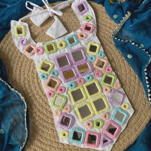 Può includere: Bavaglino in tessuto bianco ornato da specchi quadrati colorati e dettagli all'uncinetto. Il bavaglino presenta una chiusura con fiocco ed è decorato con accenti all'uncinetto e specchio dai colori pastello. Il design è ambientato su uno sfondo neutro intrecciato.