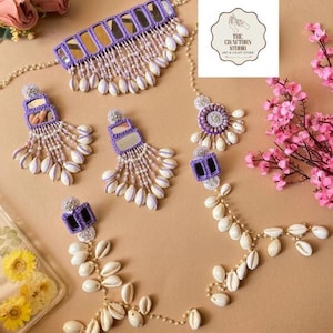 Può includere: Un set di gioielli con una collana girocollo viola con dettagli a specchio e conchiglie bianche pendenti. Sono inclusi anche orecchini e un copricapo abbinati. Il set è adornato con piccole perline e conchiglie bianche aggiuntive.