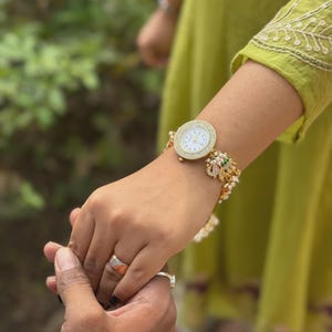 May include: Gold-toned watch with a white face and crystal-studded bezel. The watch is adorned with pearls and colourful beads. A person is wearing the watch and holding another person's hand, both wearing rings.