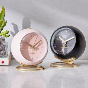 May include: Two spherical quartz clocks. One is light pink with gold hands and a gold base. The other is black with gold hands and a gold base. Both clocks have the word "QUARTZ" printed on the clock face.