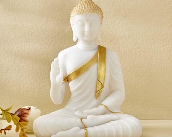 May include: A white Buddha statue in a seated meditation pose. The statue has a gold-colored textured crown and a gold sash draped over the shoulder. The statue is made of a smooth, white material.