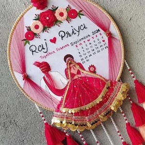 Könnte beinhalten: Runder, bestickter Wandbehang mit den Namen "Raj" und "Priya", einem Herzsymbol und dem Text "Together Forever September 2024". Enthält einen Kalender und Illustrationen eines Paares in traditioneller Kleidung. Verziert mit roten Quasten und floralen Akzenten.