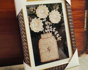Könnte beinhalten: Gerahmtes Kunstwerk mit einem Blumenarrangement aus Muscheln. Die Blumen und die Vase bestehen aus cremefarbenen Muscheln, vor einem dunklen Hintergrund. Der Rahmen hat ein dekoratives Muster in Braun und Gold, mit einem weißen Passepartout.