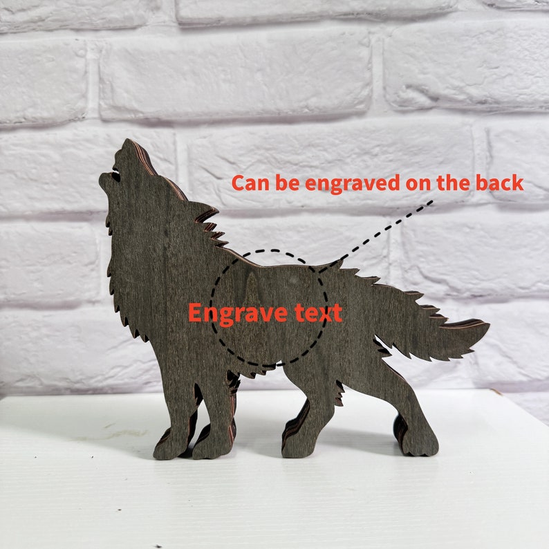 May include: A wooden wolf silhouette with a grey woodgrain finish. The wolf is howling and has a space on the back for engraving text. "Can be engraved on the back" and "Engrave text" are written on the image.
