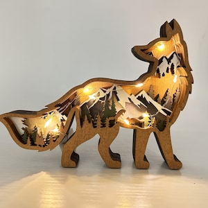 May include: A wooden fox silhouette with a layered mountain scene inside. The fox is standing on a white surface and has warm white LED lights inside.