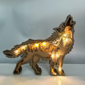 May include: A wooden wolf silhouette with a forest scene inside, illuminated by warm white fairy lights. The wolf is howling at the moon, and the forest scene includes trees, mountains, and a small wolf.