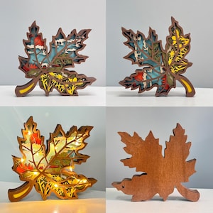 Puede incluir: Un conjunto de cuatro decoraciones de hojas de arce de madera. Tres hojas son multicapa con escenas de paisajes coloridos, mientras que una es marrón sólido. Una hoja tiene luces LED blancas cálidas.