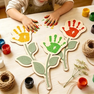 Peut inclure: Découpes de fleurs en bois avec des empreintes de mains peintes en jaune, rouge et vert. Chaque fleur a une tige et des feuilles, avec le texte "Happy Mother's Day" et une date. Des pots de peinture et de la ficelle sont dispersés.