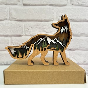 May include: Wooden fox silhouette with a mountain landscape scene inside. The fox is standing on a brown wooden base.