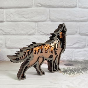May include: A wooden wolf sculpture with a layered design, featuring a forest scene inside the wolf's silhouette. The wolf is howling and the forest scene is in shades of brown, orange, and white.