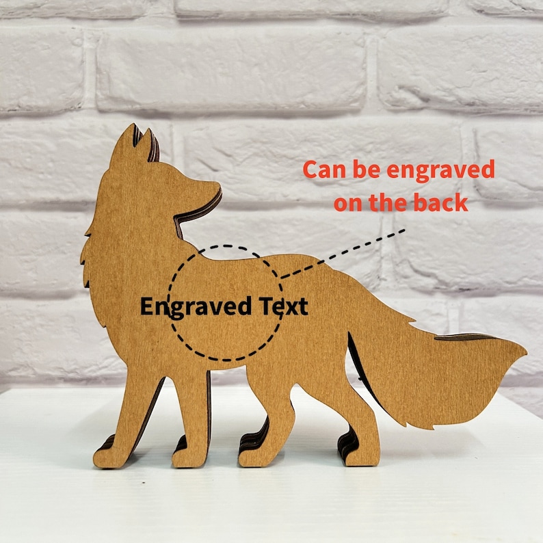 May include: Wooden cutout of a fox standing on a white surface. The fox is brown and has a long tail. The back of the fox can be engraved with text. "Engraved Text" is written on the fox's back.