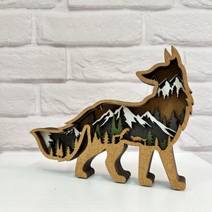 May include: A wooden fox silhouette with a layered design featuring a mountain scene with trees and snow. The fox is brown and the mountain scene is in shades of brown, green, and white.