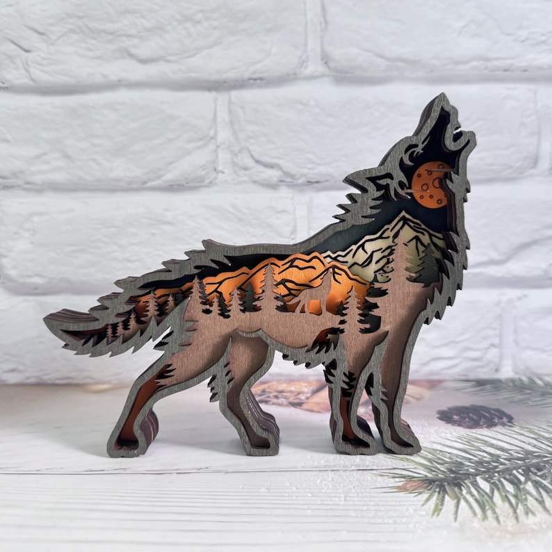 May include: A wooden wolf sculpture with a layered design, featuring a mountain range and forest scene inside the wolf's silhouette. The wolf is howling at a full moon, which is depicted in orange and brown tones.