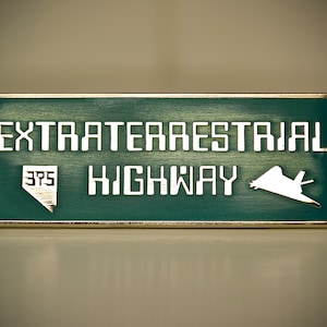 Puede incluir: Un letrero de metal verde con ribete dorado dice "Extraterrestrial Highway 375" con una silueta blanca de una nave espacial.