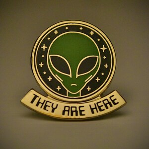 May include: Gold-coloured enamel pin with a green alien head in a black circle with gold stars and the text "They Are Here" on a gold banner.