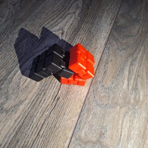 Può includere: Un giocattolo cubo fidget nero e rosso, composto da più piccoli cubi collegati tra loro. Il giocattolo è appoggiato su una superficie di legno, con la sezione rossa al centro. Il cubo è progettato per essere manipolato e giocato.
