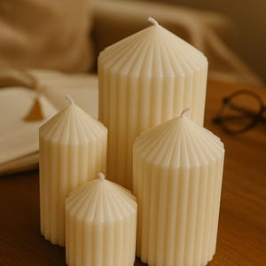 Peut inclure: Quatre bougies piliers ivoire de différentes hauteurs, disposées sur une surface en bois. Chaque bougie présente un design côtelé et un sommet conique. Les bougies ne sont pas allumées, suggérant un usage décoratif ou d'ambiance.