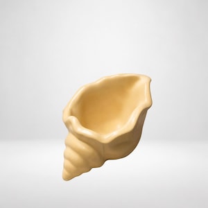 Op de afbeelding: Een lichtbeige keramische kom in de vorm van een schelp. Het object heeft een gladde, matte afwerking met een brede, open opening en een spiraalvormige, taps toelopende basis. De binnenkant is hol en concaaf, gelijk aan natuurlijke schelpvormen.