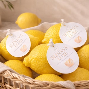 Pode incluir: Uma cesta de vime cheia de limões amarelos brilhantes, alguns com etiquetas brancas. As etiquetas dizem "SLINNVA LUXE" em letras douradas. Um vaso branco e limões adicionais estão no fundo.