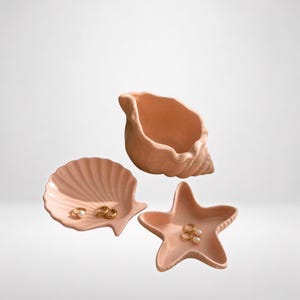 Op de afbeelding: Een set van drie licht perzikkleurige keramische schaaltjes in de vorm van zeedieren, waaronder een schelpvormige kom, een sint-jakobsschelp-schaaltje en een zeester-schaaltje. De schaaltjes bevatten kleine gouden ringen met witte parels.