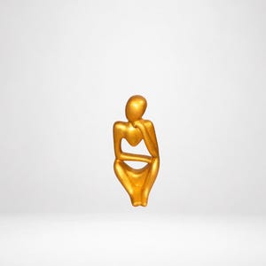 Op de afbeelding: Een goudkleurig beeldhouwwerk van een zittende figuur in een contemplatieve houding. De figuur heeft een glad, afgerond hoofd, één arm rustend tegen de kin en benen naar binnen gevouwen, wat een doorlopend, vloeiend silhouet met een metallic afwerking creëert.