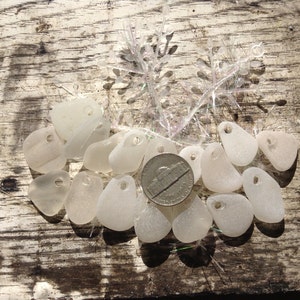 Puede incluir: Una colecci&oacute;n de amuletos de vidrio de mar blanco con un agujero perforado en cada pieza. Los amuletos est&aacute;n dispuestos sobre una superficie de madera con una moneda de plata en el centro.