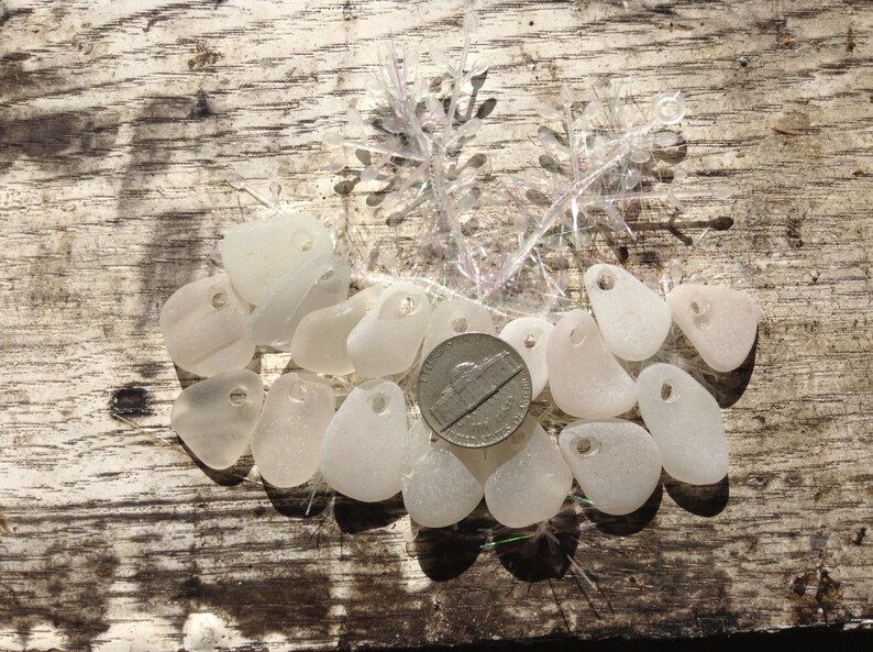 Puede incluir: Una colecci&oacute;n de amuletos de vidrio de mar blanco con un agujero perforado en cada pieza. Los amuletos est&aacute;n dispuestos sobre una superficie de madera con una moneda de plata en el centro.