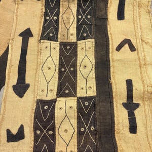 May include: A textile wall hanging featuring a patchwork of beige and dark brown fabric panels. The beige sections display black geometric symbols, including arrows and crosses, while the dark brown panels are decorated with white stitched diamond patterns and small circular motifs.