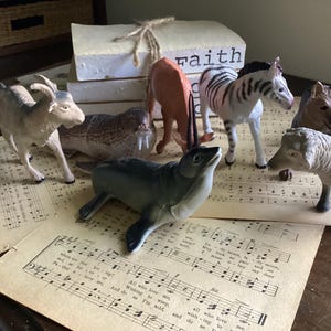 Puede incluir: Una colección de figuritas de animales de plástico, que incluyen una cabra, una morsa, una foca, una cebra, una llama, un lobo y una oveja. Las figuritas están dispuestas sobre una hoja de música con la palabra "fe" visible en una pila de libros en el fondo.
