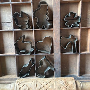 May include: A collection of eight metal biscuit cutters in various shapes including a bird, a person, a flower, a rabbit, a heart, an axe, and two abstract forms, arranged in a wooden printer's tray above a carved wooden rolling pin.