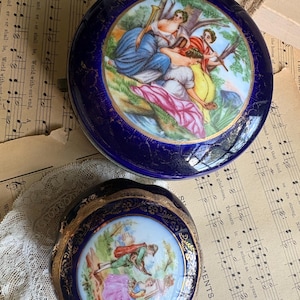 May include: Two round porcelain trinket boxes with deep blue borders and gold filigree accents. The larger box features a painted scene of three figures in a landscape, while the smaller box depicts two figures in a garden setting with colourful clothing.
