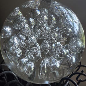 May include: A clear glass paperweight with numerous internal bubbles, creating a textured, refractive effect. The spherical object sits atop a dark, wrought-iron decorative stand, adding a touch of elegance to any desk or shelf.