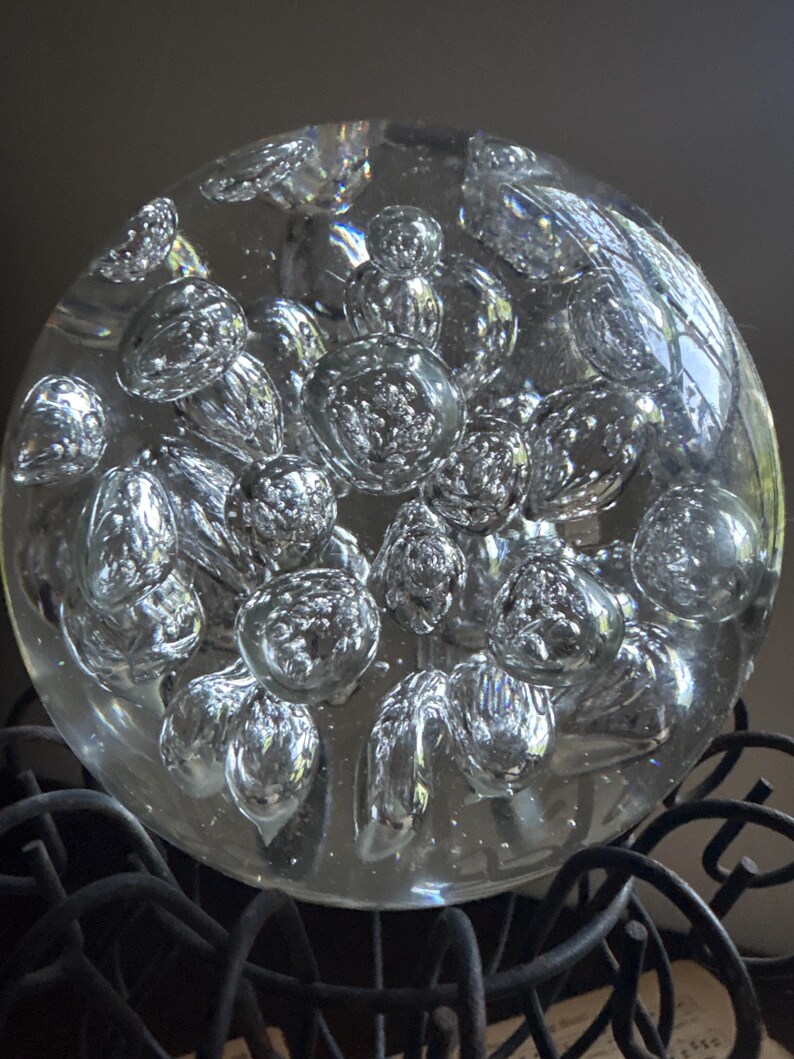 May include: A clear glass paperweight with numerous internal bubbles, creating a textured, refractive effect. The spherical object sits atop a dark, wrought-iron decorative stand, adding a touch of elegance to any desk or shelf.
