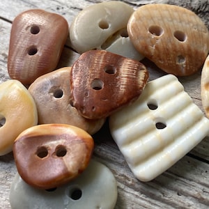 May include: A collection of natural-looking buttons in various shapes and colors, including brown, tan, and off-white. Each button has two or four holes for sewing. The buttons have a rustic, organic aesthetic.