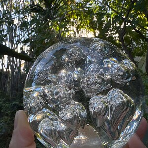May include: A clear glass sphere filled with numerous air bubbles. The sphere is held in a hand, with a blurred background of green foliage and sunlight. The bubbles are reflective, creating a shimmering effect.