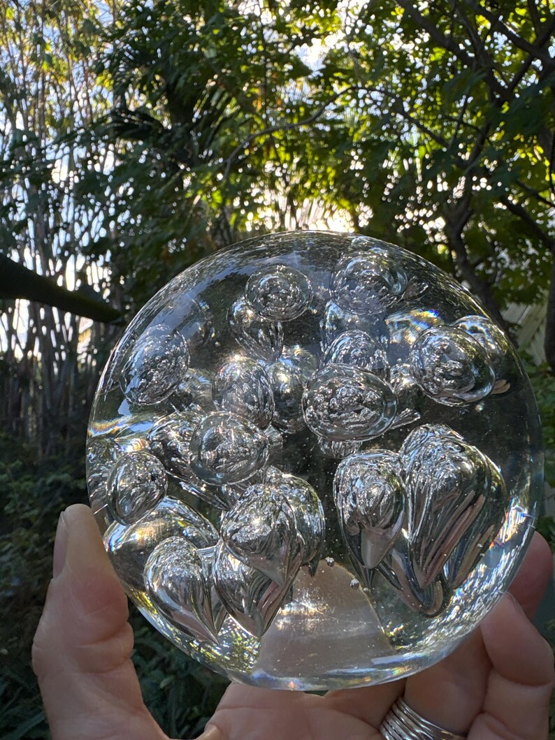 May include: A clear glass sphere filled with numerous air bubbles. The sphere is held in a hand, with a blurred background of green foliage and sunlight. The bubbles are reflective, creating a shimmering effect.