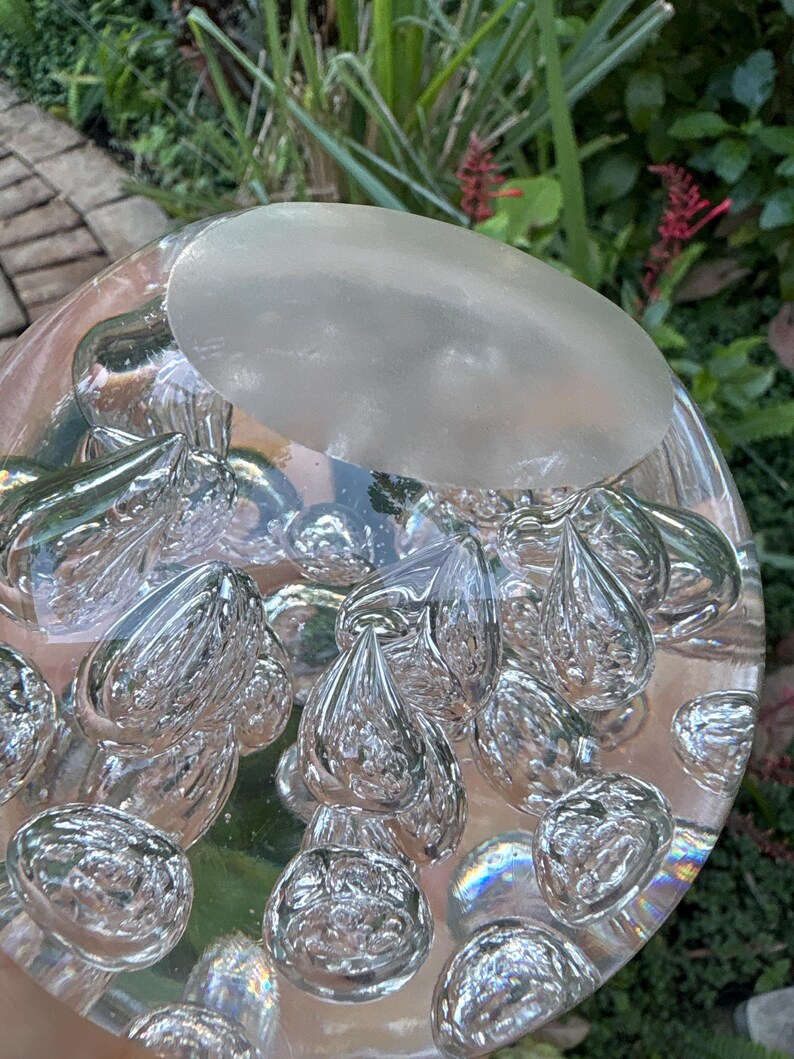 May include: A clear glass paperweight with a frosted oval top and numerous teardrop-shaped bubbles inside. The bubbles are reflective, and the paperweight sits on a reflective surface. The background features green foliage.