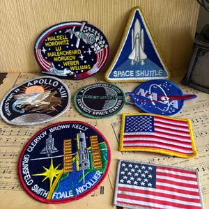 Peut inclure: Une collection d'écussons brodés sur le thème de l'espace, de formes circulaires, triangulaires et rectangulaires. Les motifs incluent des logos de la NASA, la navette spatiale, Apollo XIII, le Kennedy Space Center et deux drapeaux américains.