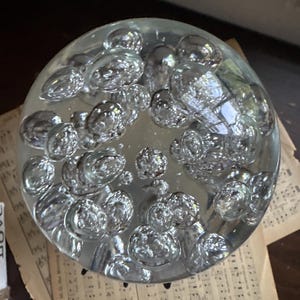 May include: A clear glass paperweight with a bubble design. The round paperweight is filled with numerous spherical air bubbles, creating a textured appearance. The paperweight sits on top of vintage sheet music.